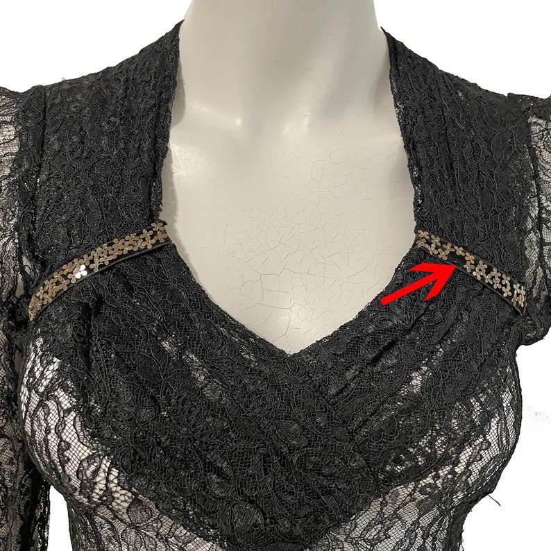 K&ouml;nnte beinhalten: Schwarzes Spitzentop mit langen &Auml;rmeln und V-Ausschnitt. Das Oberteil hat ein zartes Blumenmuster und ist mit einem dekorativen Paillettenband am Ausschnitt verziert. Das Kleidungsst&uuml;ck wird auf einer Schaufensterpuppe pr&auml;sentiert.