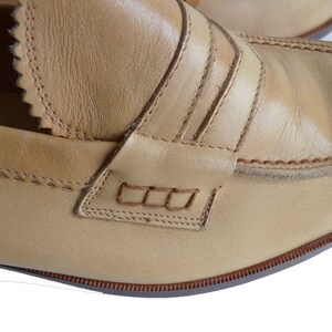 Puede incluir: Primer plano de un par de mocasines penny de cuero color canela. Los zapatos presentan costuras decorativas, un cuello con muescas y un dise&ntilde;o cl&aacute;sico de ranura para penique. Las suelas son de color marr&oacute;n contrastante.