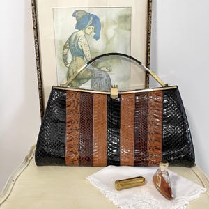 May include: A vintage handbag with a black, brown, and tan snakeskin pattern. The bag has a gold-tone clasp and handle. A framed artwork and a small perfume bottle are in the background.
