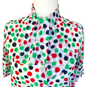 May include: A white blouse with a large green, red, and black polka dot pattern. The blouse has a large, tied bow at the neck.