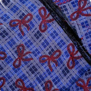 May include: A blue fabric featuring a dark blue plaid pattern and embroidered red glittery bow motifs. The material has a sheer, textured quality with a distinct black fringed edge running vertically along the side of the textile piece.