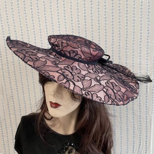 Peut inclure: Un chapeau à larges bords avec une superposition de dentelle florale rose foncé et noire. Le chapeau présente une couronne arrondie et un large bord, avec un ruban noir et des détails de plumes. Le chapeau est présenté sur un mannequin.
