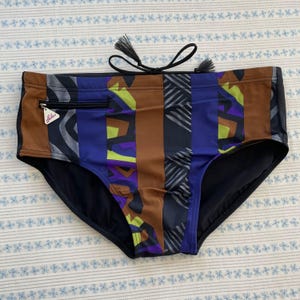 Pode incluir: Calções de banho com um padrão listrado colorido. Os calções de banho apresentam uma cintura preta e um cordão com borlas. A frente dos calções de banho tem um bolso com zíper e uma variedade de cores, incluindo castanho, azul e roxo.