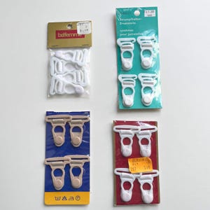 Könnte beinhalten: Gezeigt werden vier Packungen mit Dessous-Zubehör. Zwei Packungen enthalten weiße Strumpfhalter-Clips, eine mit goldenem Etikett. Die anderen beiden Packungen enthalten beige und weiße Strumpfhalter-Clips, jeweils auf farbigem Träger.