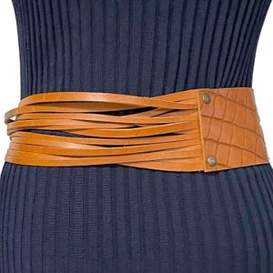 May include: A tan leather belt with a wide, textured crocodile-style panel and multiple thin, layered straps. The belt is secured with two metal rivets and is shown against a dark blue ribbed fabric.