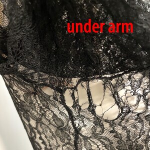 K&ouml;nnte beinhalten: Nahaufnahme von schwarzem Spitzenstoff mit den roten Worten "under arm". Die Spitze hat ein zartes, offenes Webmuster, das einen durchsichtigen Effekt erzeugt. Der Stoff scheint Teil eines Kleidungsst&uuml;cks zu sein.