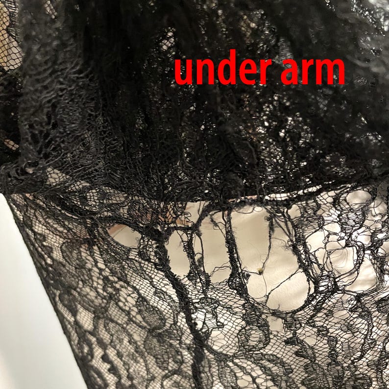 K&ouml;nnte beinhalten: Nahaufnahme von schwarzem Spitzenstoff mit den roten Worten "under arm". Die Spitze hat ein zartes, offenes Webmuster, das einen durchsichtigen Effekt erzeugt. Der Stoff scheint Teil eines Kleidungsst&uuml;cks zu sein.