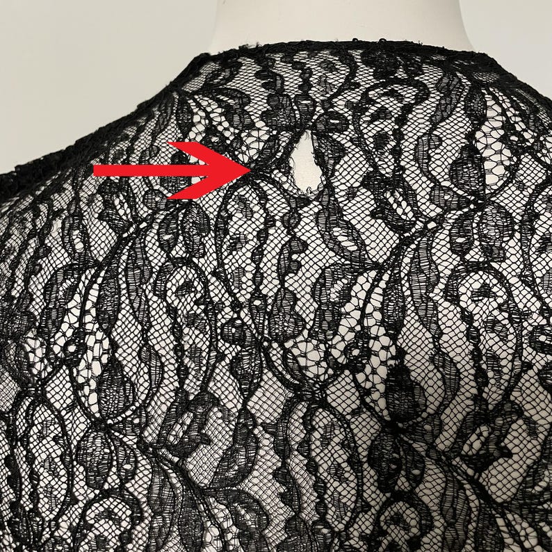 K&ouml;nnte beinhalten: Nahaufnahme eines schwarzen Spitzengewebes mit floralem Muster. Der zarte Stoff ist &uuml;ber einer wei&szlig;en Oberfl&auml;che drapiert, wodurch die komplizierten Details der Spitze hervorgehoben werden. Ein kleines Loch ist in der Spitze sichtbar.
