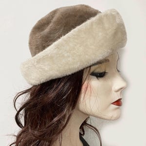 May include: A cloche-style hat made of faux fur. The hat has a light brown crown and a broad, cream-coloured brim. The hat is designed to be worn on the head and is shown on a mannequin.