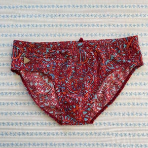 May include: Red and blue patterned underwear. The underwear features a repeating pattern of abstract shapes in red, orange, and blue. The edges are trimmed in a darker red. A small, triangular tag is visible on the front.