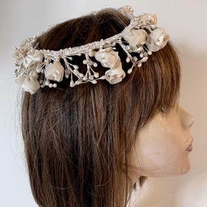 May include: A white floral crown with pearl and crystal accents. The crown is made of silk flowers and is designed to be worn on the head.