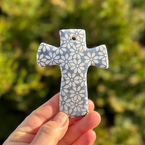May include: A ceramic cross with a blue and white floral pattern. The cross has a hole at the top for hanging. The design features small white flowers on a blue background. The cross is held up against a blurred green background.