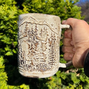 May include: A beige ceramic mug with a detailed forest scene carved into the surface. The design includes trees, mountains, and stars. The mug is held against a backdrop of green foliage.