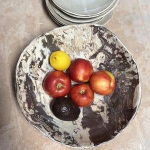 May include: A large, decorative ceramic bowl with a marbled brown and cream pattern, filled with fresh fruit. The bowl contains red apples, a yellow lemon, and a dark avocado. A stack of matching plates is visible in the background.