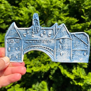 May include: A blue and white ceramic ornament shaped like a building with a bridge. The ornament has a textured surface and is held in a hand against a backdrop of green foliage. The building features architectural details.