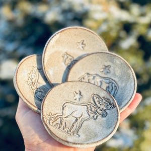 May include: A set of four small, round ceramic plates. Each plate features a different embossed design of a moose, a bear, a plant, and a star. The plates are a neutral beige color with a slightly textured surface.
