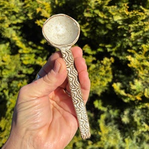 May include: A handmade ceramic spoon with a speckled off-white bowl and a handle decorated with a brown wave pattern. The spoon is held against a backdrop of green foliage, showcasing its rustic charm.