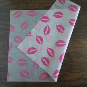 May include: Two sheets of tissue paper with a repeating pattern of pink lips. The paper is semi-transparent, with the pink lips printed across the surface. One sheet is partially folded over the other, revealing the pattern.