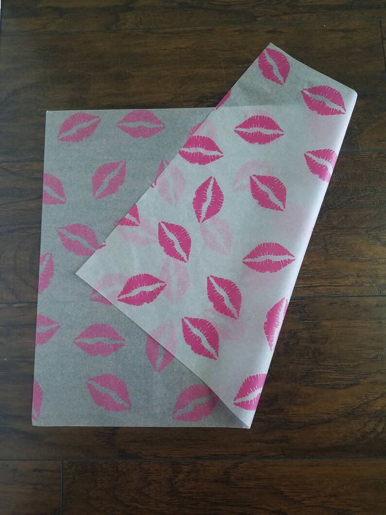 May include: Two sheets of tissue paper with a repeating pattern of pink lips. The paper is semi-transparent, with the pink lips printed across the surface. One sheet is partially folded over the other, revealing the pattern.