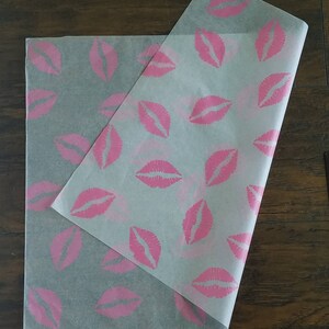 May include: Two sheets of translucent paper with a repeating pattern of pink lips. One sheet is folded over the other, revealing the design. The paper is likely used for wrapping or packaging, with a playful and feminine theme.