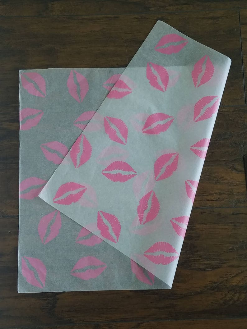 May include: Two sheets of translucent paper with a repeating pattern of pink lips. One sheet is folded over the other, revealing the design. The paper is likely used for wrapping or packaging, with a playful and feminine theme.