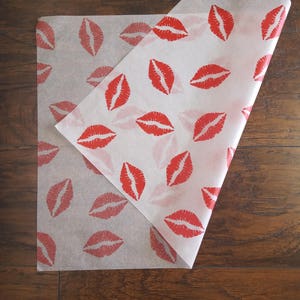 May include: White tissue paper with a repeating pattern of red lipstick kiss prints. The paper is folded over, revealing the pattern on both sides. The paper is on a dark wood surface.