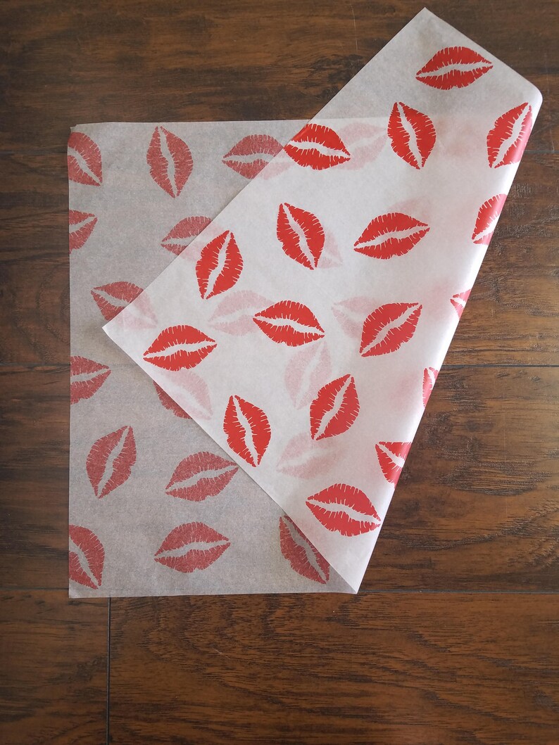 May include: White tissue paper with a repeating pattern of red lipstick kiss prints. The paper is folded over, revealing the pattern on both sides. The paper is on a dark wood surface.