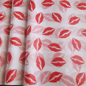 May include: White wrapping paper with a repeating pattern of red lipstick kiss prints. The paper is folded, showing multiple layers of the printed design. The kisses are in a bold red color, with a lighter red shadow effect.