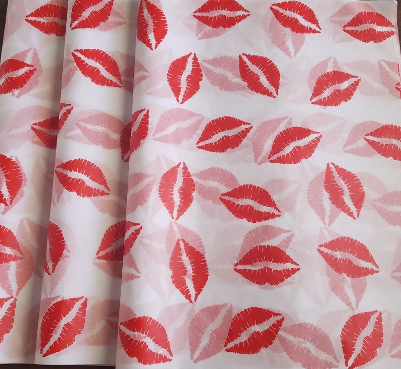 May include: White wrapping paper with a repeating pattern of red lipstick kiss prints. The paper is folded, showing multiple layers of the printed design. The kisses are in a bold red color, with a lighter red shadow effect.