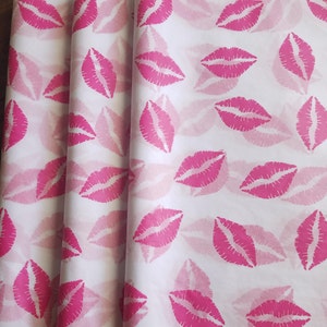 May include: White wrapping paper with a repeating pattern of bright pink lips. The lips are stylized and evenly spaced across the paper, creating a playful and feminine design. The paper is folded, showing the texture and layers.