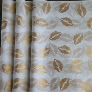 May include: White wrapping paper with a repeating pattern of gold lips. The lips are in a kiss shape and are arranged in a grid-like fashion. The paper is rolled up in a stack.
