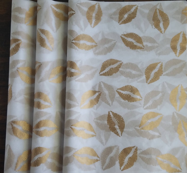 May include: White wrapping paper with a repeating pattern of gold lips. The lips are in a kiss shape and are arranged in a grid-like fashion. The paper is rolled up in a stack.