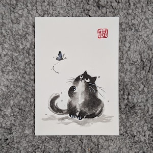 以下が含まれることがあります： 白い紙に描かれた長方形のアートプリントで、小さな青い蝶を見上げるふわふわした猫の黒い墨絵が特徴です。猫は大きな目と曲がった尻尾をしています。右上隅には、漢字が刻まれた赤い四角いスタンプが押されています。