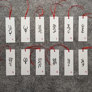 May include: A collection of white rectangular bookmarks with black calligraphy and red ribbon ties. Each bookmark features a unique abstract design and a small red stamp. The bookmarks are arranged on a gray surface.