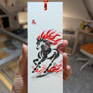 May include: A white bookmark featuring a watercolour painting of a black horse with a red mane and flames. The horse is running. A red ribbon is attached at the top. Chinese characters are at the top.