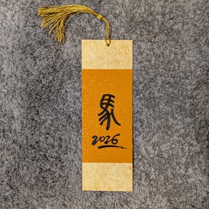 Könnte beinhalten: Rechteckiger Lesezeichen mit goldener Quaste. Das Lesezeichen hat eine hellbeige Ober- und Unterseite mit einem orangefarbenen Mittelteil. Schwarze Kalligraphie und die Zahl 2026 sind auf dem orangefarbenen Abschnitt aufgedruckt.
