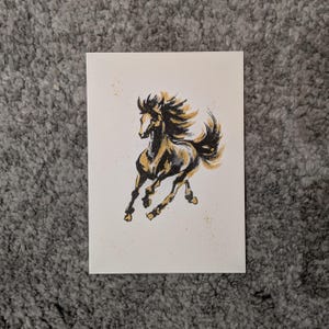 Könnte beinhalten: Ein handgemaltes Kunstwerk mit einem galoppierenden Pferd. Das Pferd ist in Schwarz- und Goldtönen dargestellt, mit fließender Mähne und Schweif. Das Kunstwerk befindet sich auf cremefarbenem Papier mit dezenten Farbspritzern.