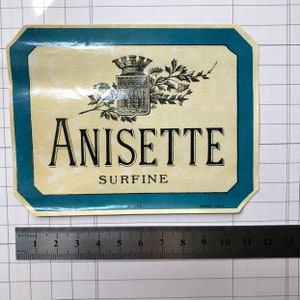 Op de afbeelding: Een vintage etiket voor Anisette Surfine, een Franse likeur. Het etiket heeft een wapenschild met een kroon en een stadsbeeld, omringd door een bloemmotief. De tekst "Anisette Surfine" is gedrukt in zwarte letters op een crèmekleurige achtergrond, met een blauwe rand.