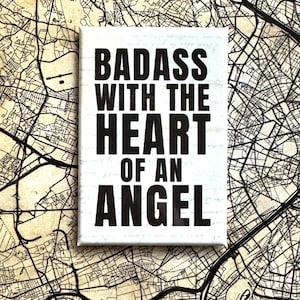 Op de afbeelding: Een rechthoekig wit canvas met de tekst "BADASS WITH THE HEART OF AN ANGEL" in dikke zwarte letters. Het canvas is geplaatst tegen een vintage kaartachtergrond, wat een contrast creëert tussen de moderne tekst en de verouderde kaart.