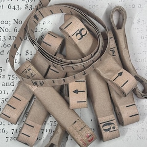 Op de afbeelding: Een verzameling beige papieren meetlinten met zwart gedrukte cijfers en markeringen. De linten zijn opgerold en gelaagd en tonen verschillende numerieke stappen en richtingpijlen die in donkere inkt op het papieroppervlak zijn gedrukt.
