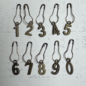 May include: A collection of ten antique bronze-colored safety pins, each with a different number from 0 to 9. The numbers are in a serif font and are suspended from the safety pin's loop.