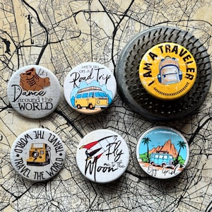 Op de afbeelding: Een verzameling ronde knopen met een reis-thema, met diverse ontwerpen en tekst. Eén knoop zegt "I AM A TRAVELER" met een rugzak illustratie. Andere knopen bevatten zinnen als "Dance around the World", "Road Trip" en "Fly me to the Moon".