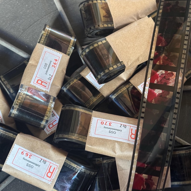 May include: A close-up of several rolls of film, wrapped in brown paper, with labels that include the text "OSS", "Zaltbommelseweg", "Os Z", and numbers. The film is black and white with a red stripe.