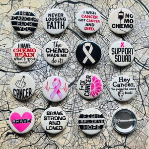 Può includere: Una collezione di spille rotonde con vari testi e design grafici. Le spille presentano frasi come "Hey Cancer Fuck You", "Never Loosing Faith" e "Support Squad". Alcune spille includono nastri rosa per il cancro e disegni floreali.