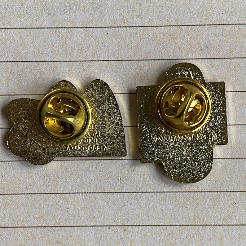May include: Two gold-colored metal pins. The first pin is shaped like the McDonald's golden arches logo. The back of the pin says "McDonald's 1998-2000 TM". The second pin is shaped like a square with rounded corners. The back of the pin says "&copy;2000 McDonald's TM".