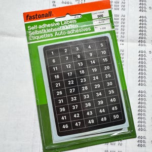 Peut inclure: Un paquet d'étiquettes autocollantes numérotées de 1 à 50, disposées en grille. Les étiquettes sont noires avec des chiffres blancs, l'emballage est vert et orange. La marque "fastonall" est visible.