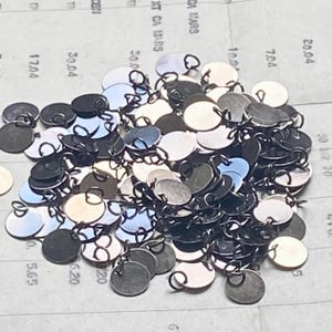 Op de afbeelding: Een stapel kleine, ronde metalen bedels met lussen. De bedels hebben verschillende kleuren, waaronder zilver, zwart en iriserende tinten. Ze liggen op een papieren oppervlak met gedrukte cijfers en tekst.
