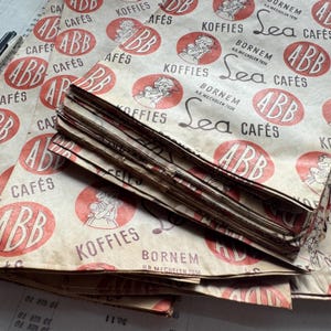 May include: A stack of vintage paper packaging with a repeating pattern of red circles, text, and illustrations. The text includes "ABB CAF&Eacute;S", "KOFFIES", and "Sea CAF&Eacute;S". The paper has a worn, aged appearance.
