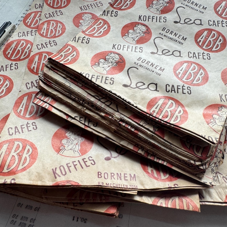 May include: A stack of vintage paper packaging with a repeating pattern of red circles, text, and illustrations. The text includes "ABB CAF&Eacute;S", "KOFFIES", and "Sea CAF&Eacute;S". The paper has a worn, aged appearance.