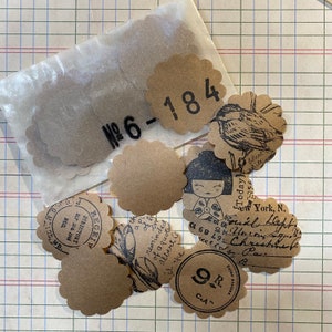 Op de afbeelding: Een set van bruine, gegolfde papieren cirkels met verschillende gedrukte ontwerpen. De ontwerpen omvatten een vogel, een Japanse pop, een bloemenpatroon en een handgeschreven notitie. De cirkels zijn genummerd "№6-184" en "9R CA".